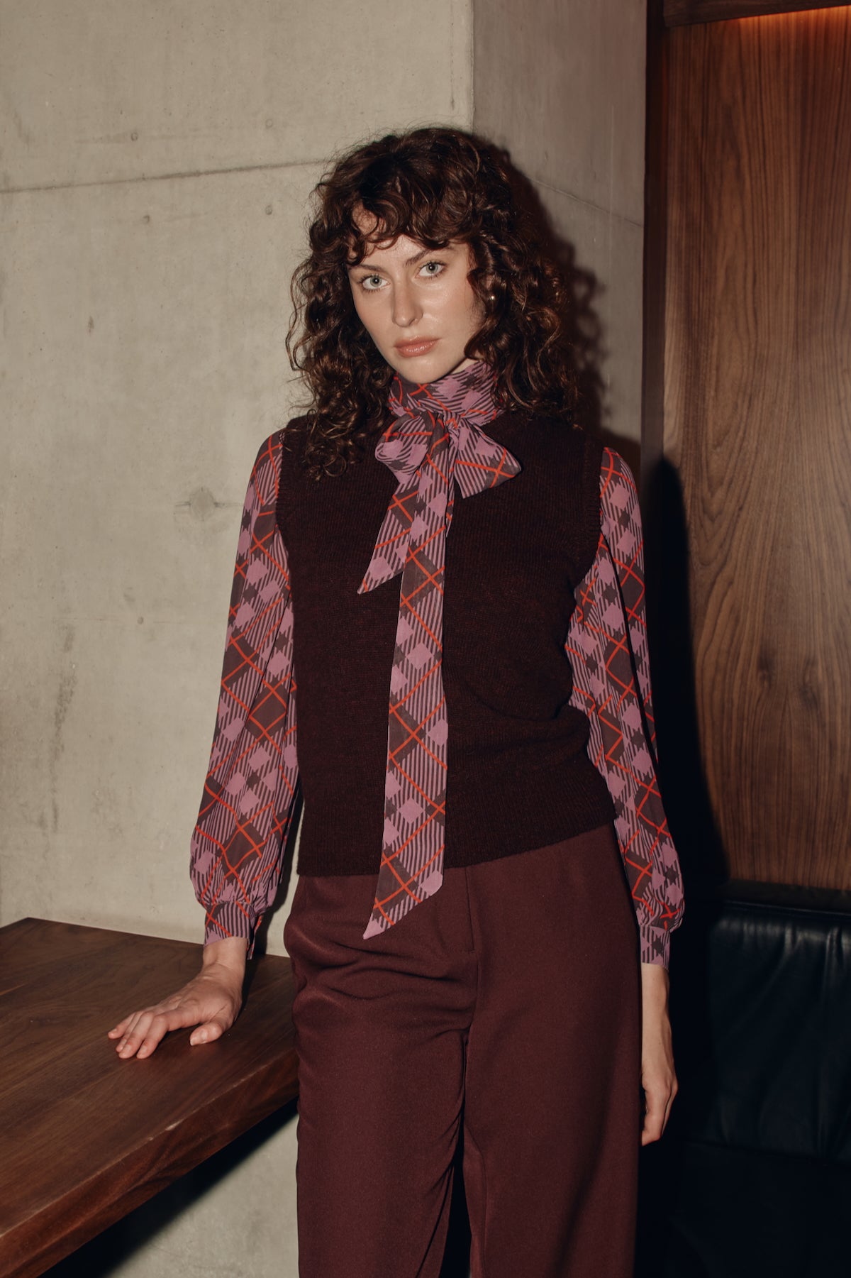Woman wearing a plaid blouse and brown outfit against a textured wall.