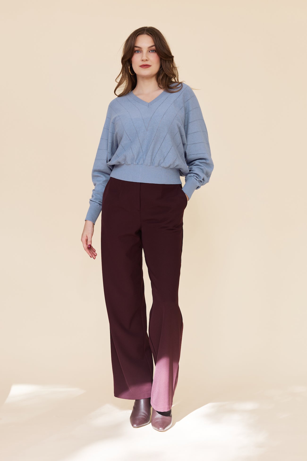 A woman modeling a light blue knit sweater and dark burgundy wide-leg trousers against a warm beige studio background.