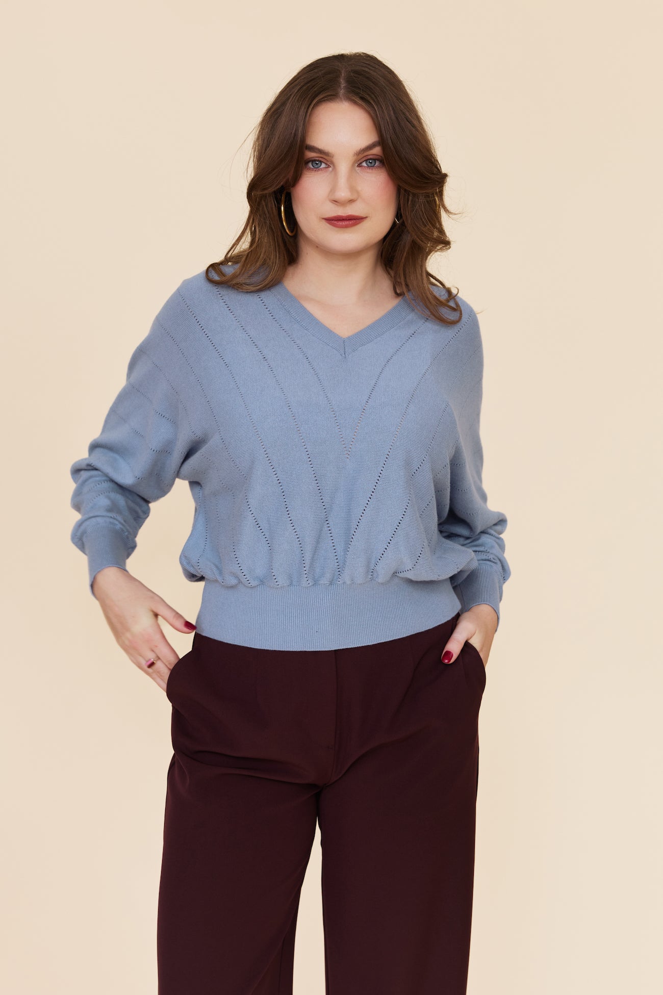 A woman modeling a light blue knit sweater with a V-neck and textured cable pattern, paired with dark burgundy pants, against a solid beige background.