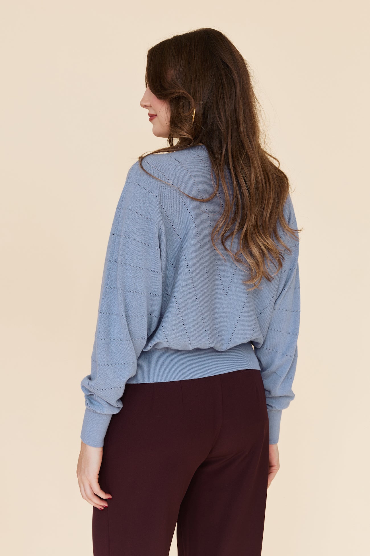A woman shown from the back wearing a light blue cropped knit sweater with a subtle grid pattern and dark burgundy pants, set against a solid beige studio background.
