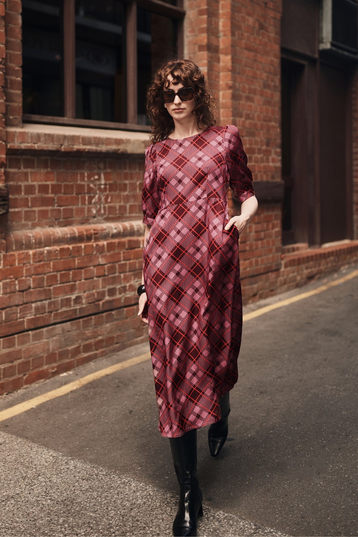 A woman wearing a burgundy and pink plaid midi dress with elbow-length sleeves walks down an outdoor street next to a brick building.