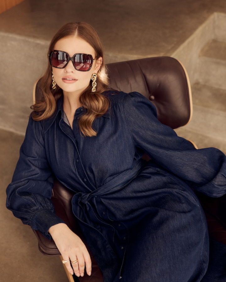 Woman in a indigo blue chambray dress sitting in a brown armchair with a beige sofa and staircase in the background.