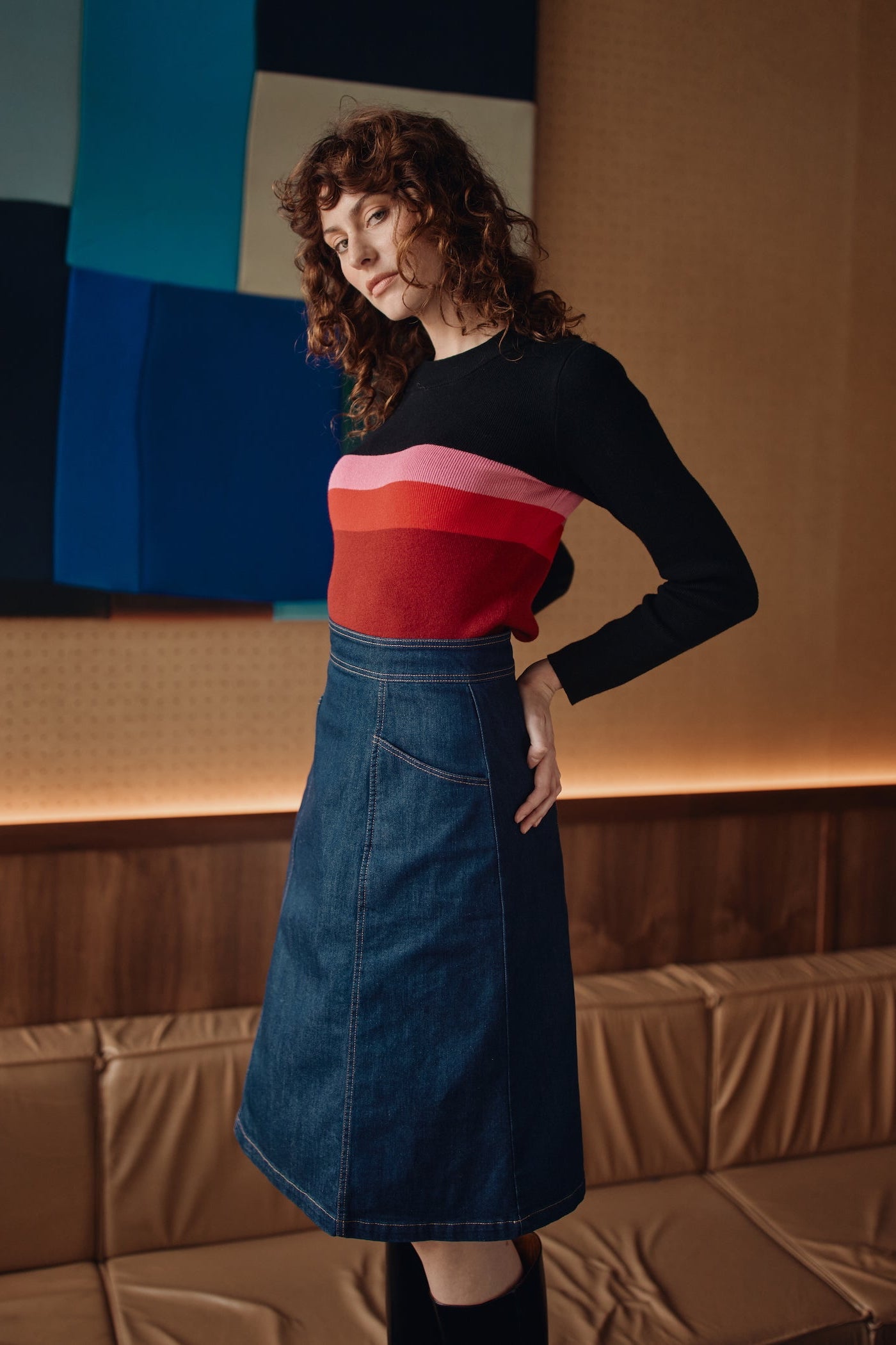 Woman wearing a colorful sweater and denim skirt in an indoor setting with geometric wall art.
