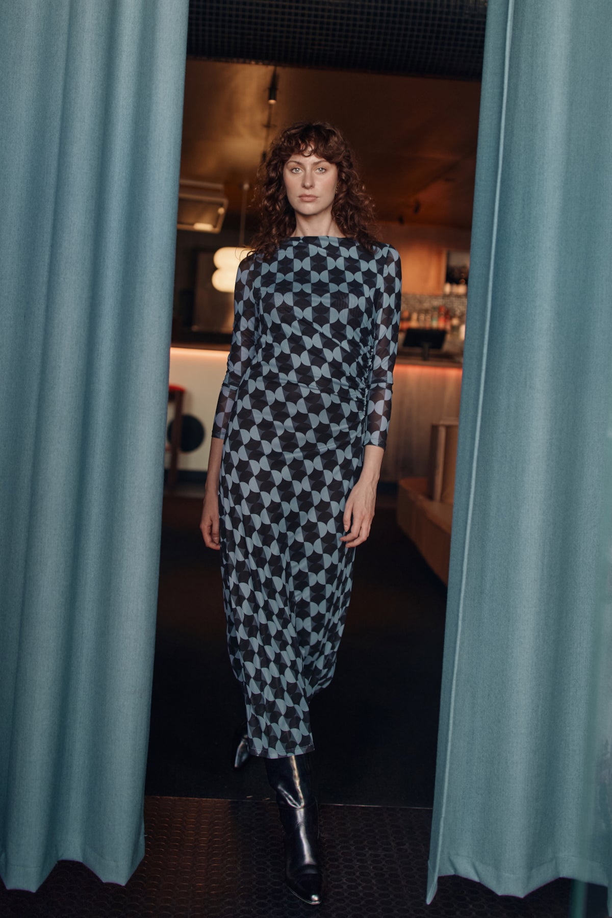 Woman in a patterned dress standing between two curtains in an indoor setting.