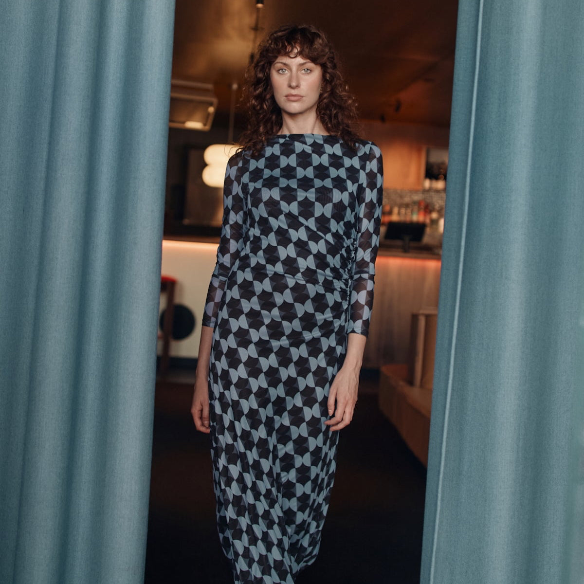 Woman in a patterned dress standing between two curtains in an indoor setting.