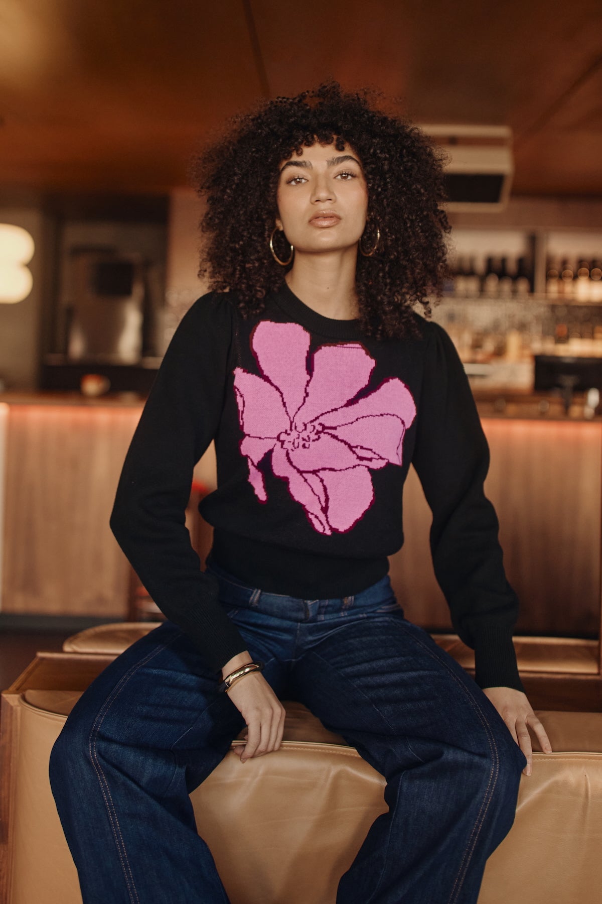Woman wearing a black sweater with a pink flower design, sitting in a bar.