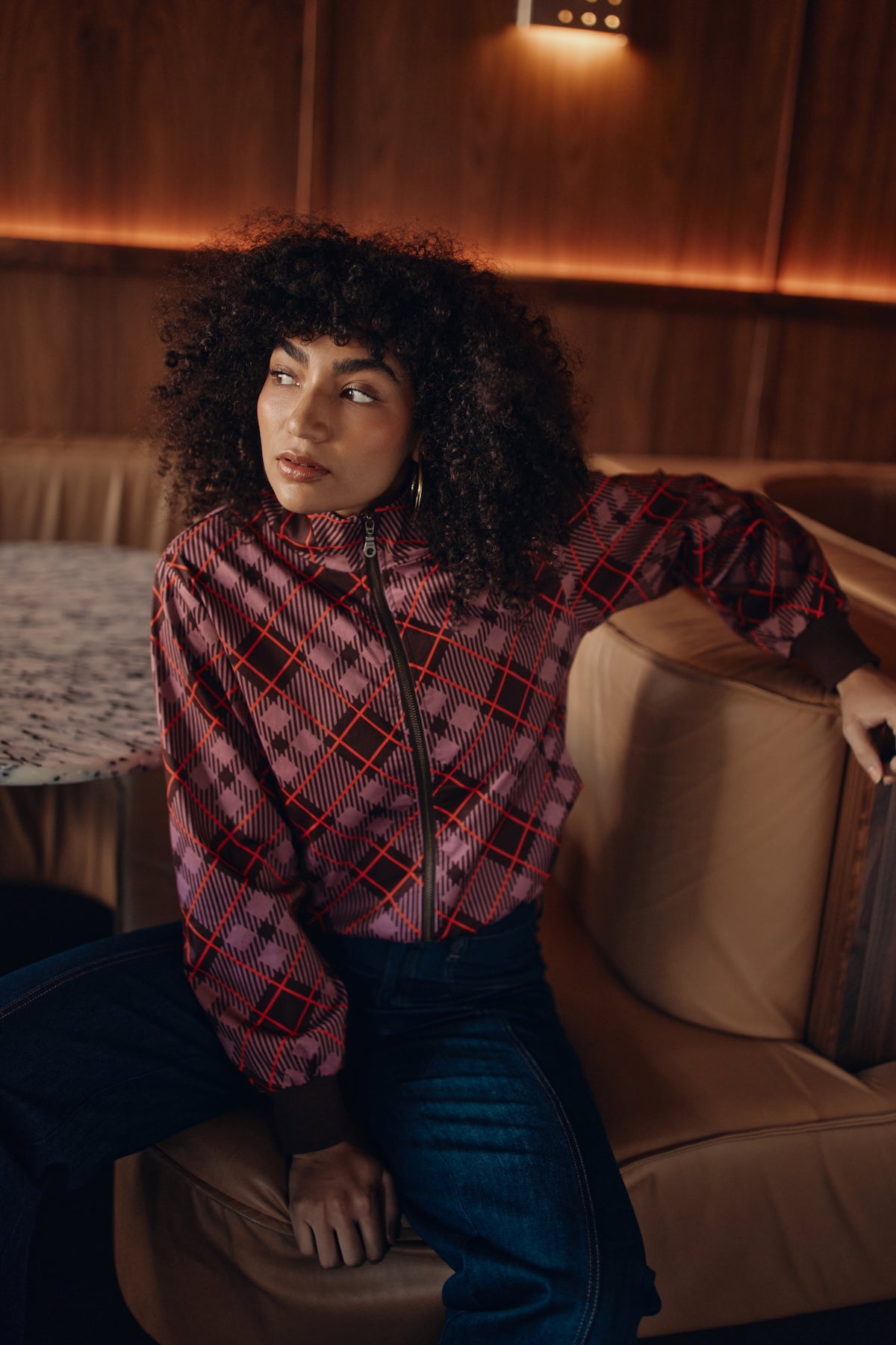 Woman in a plaid bomber jacket sitting on a couch in a warm-toned room.