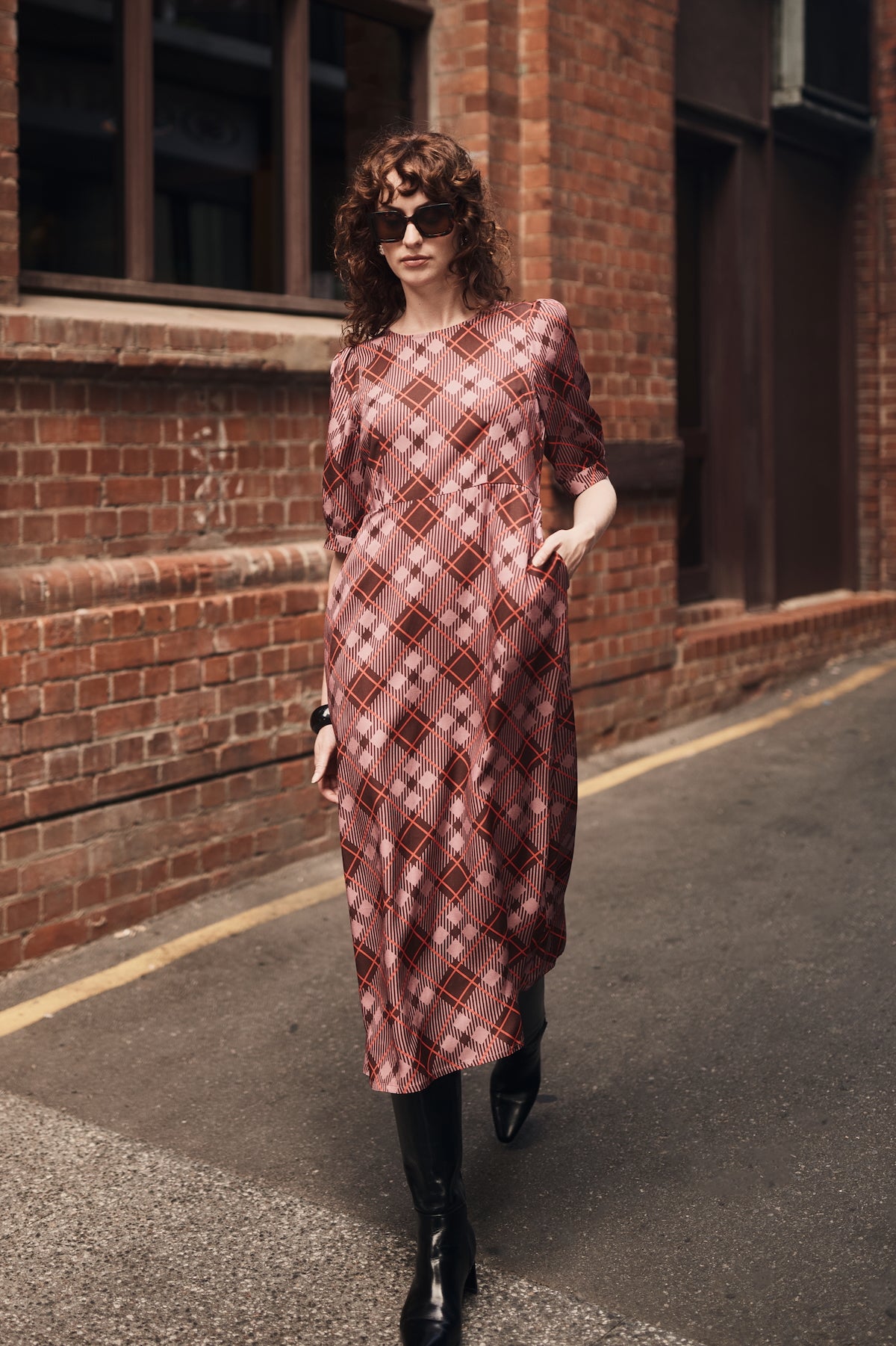 Woman in a plaid dress standing against a brick wall.