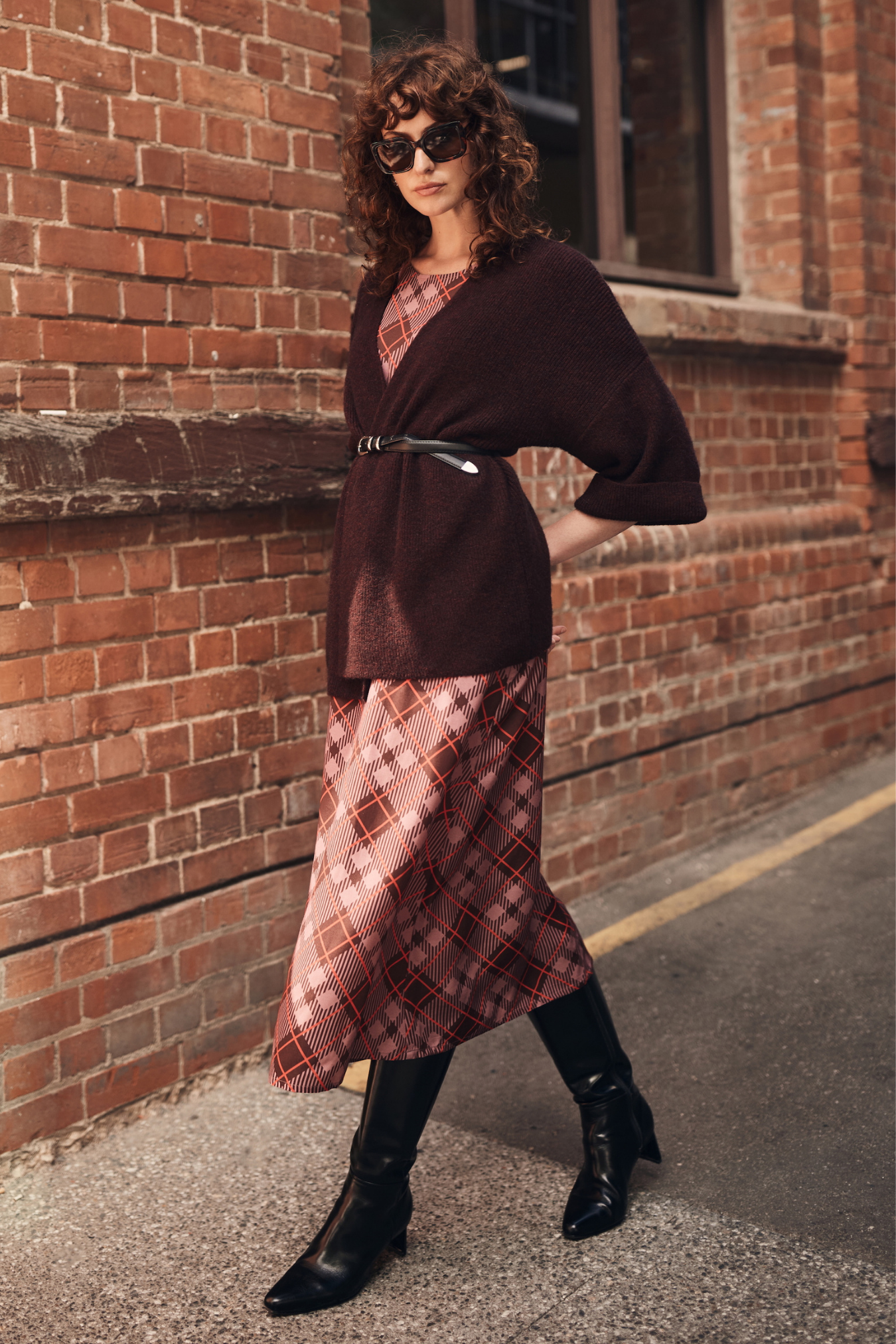 Woman in a burgundy cardigan and plaid dress standing against a brick wall.
