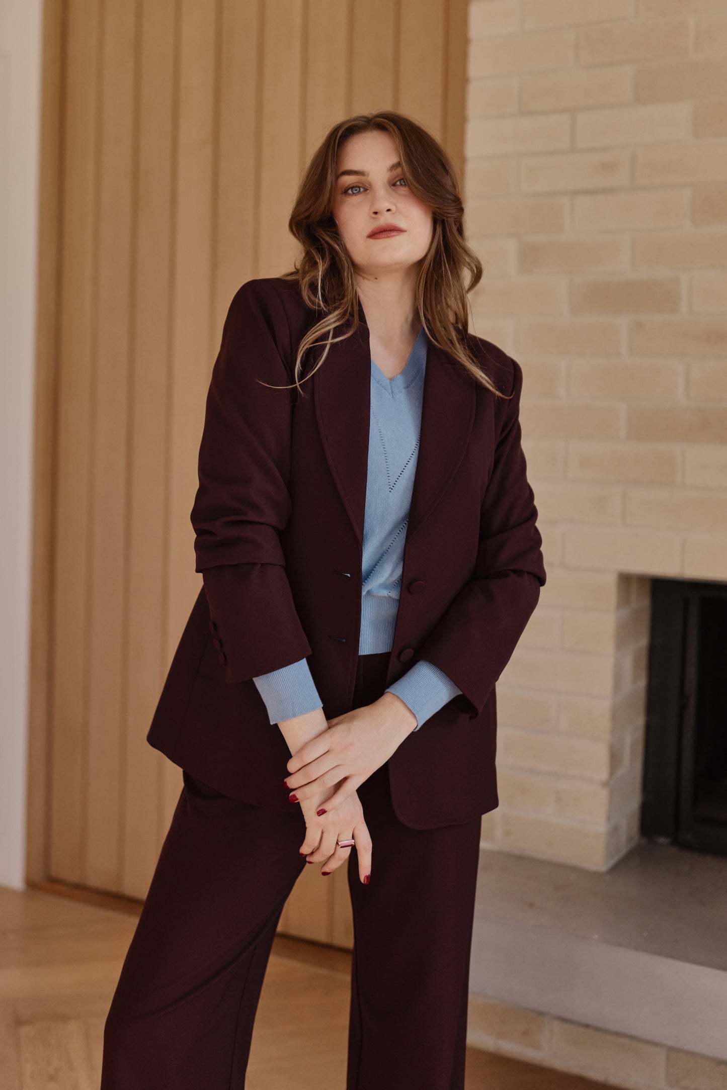 A woman models a dark burgundy two-piece suit with a light blue knitted top underneath, standing in a modern room with a wood-paneled wall and a brick fireplace.